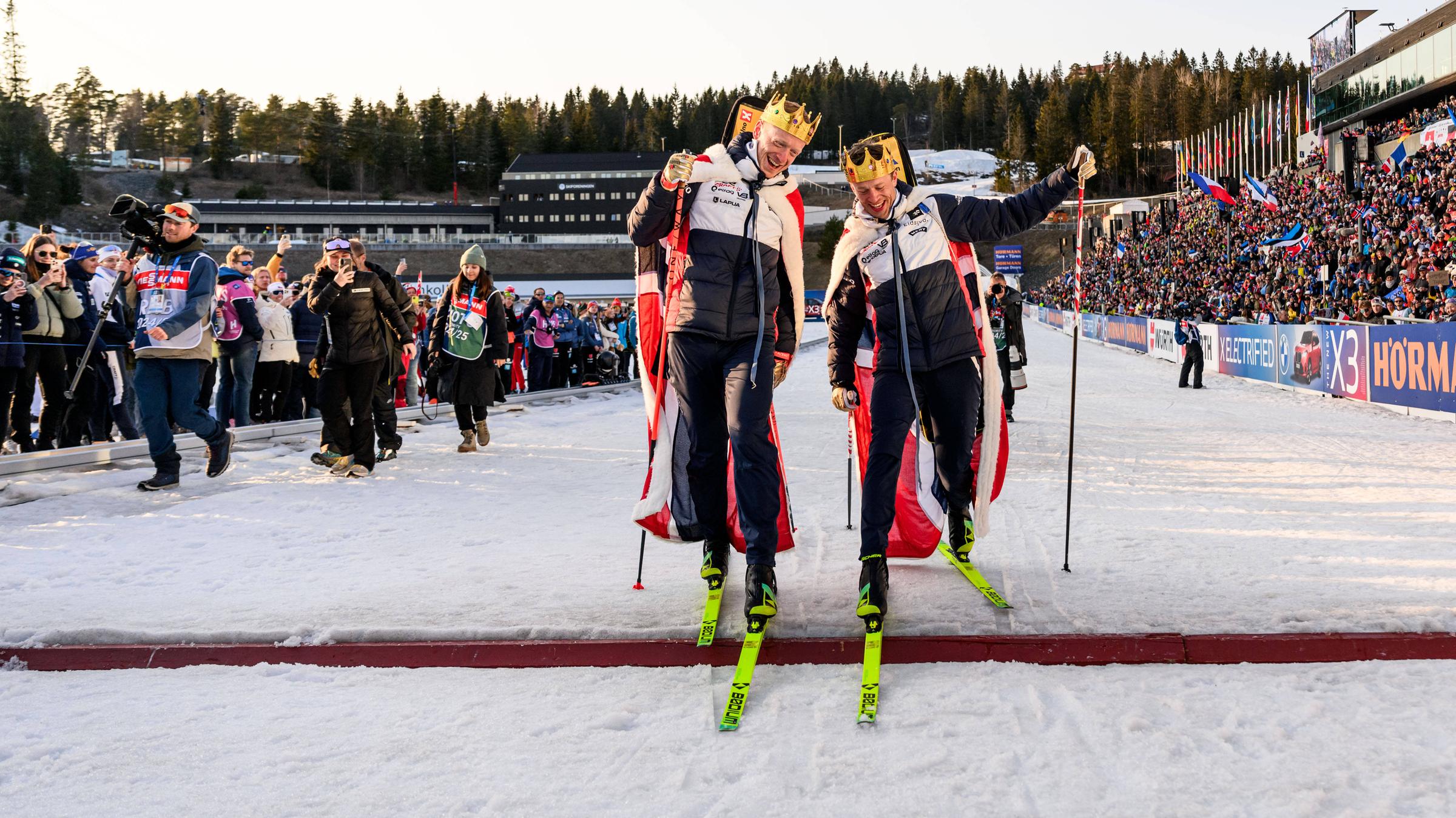 Johannes Thingnes und Tarjei Bö fahren nach dem Massenstart am 23.3.25 am Holmenkollen ein letztes Mal demonstrativ über die Ziellinie, beide mit Krone-Attrappen auf dem Kopf