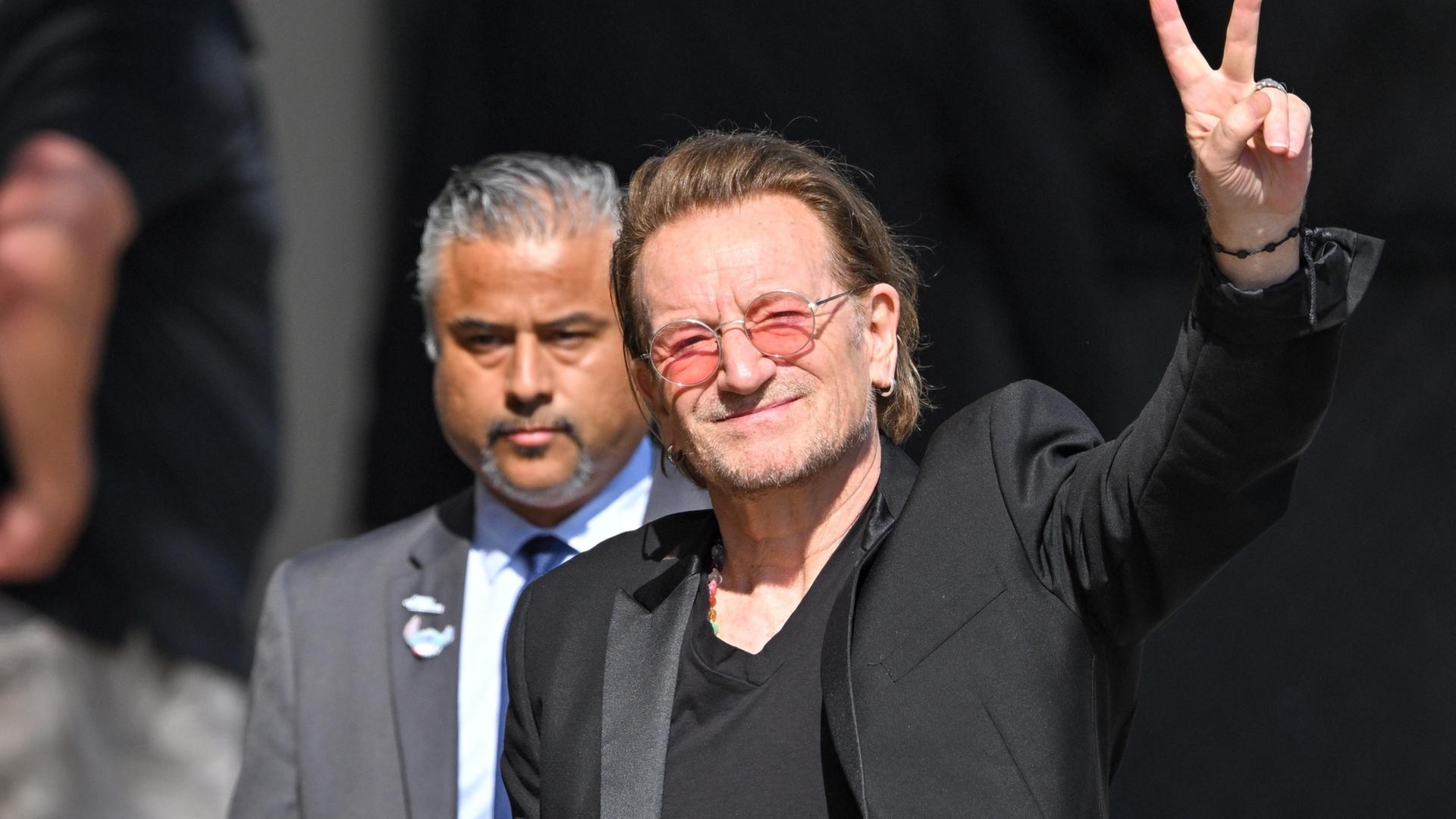 Bono lächelt und hält ein Peace-Zeichen in die Luft
