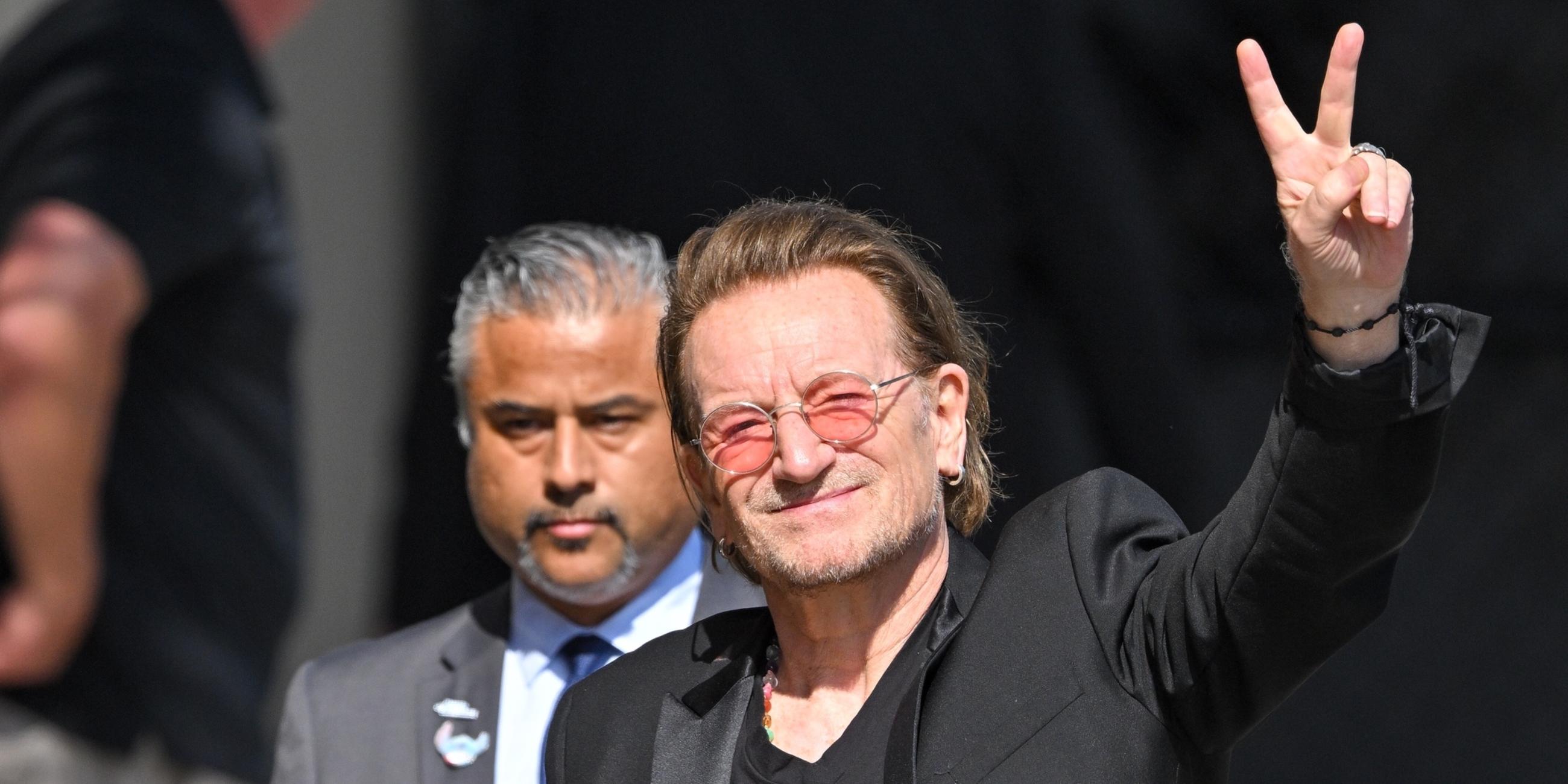 Bono lächelt und hält ein Peace-Zeichen in die Luft