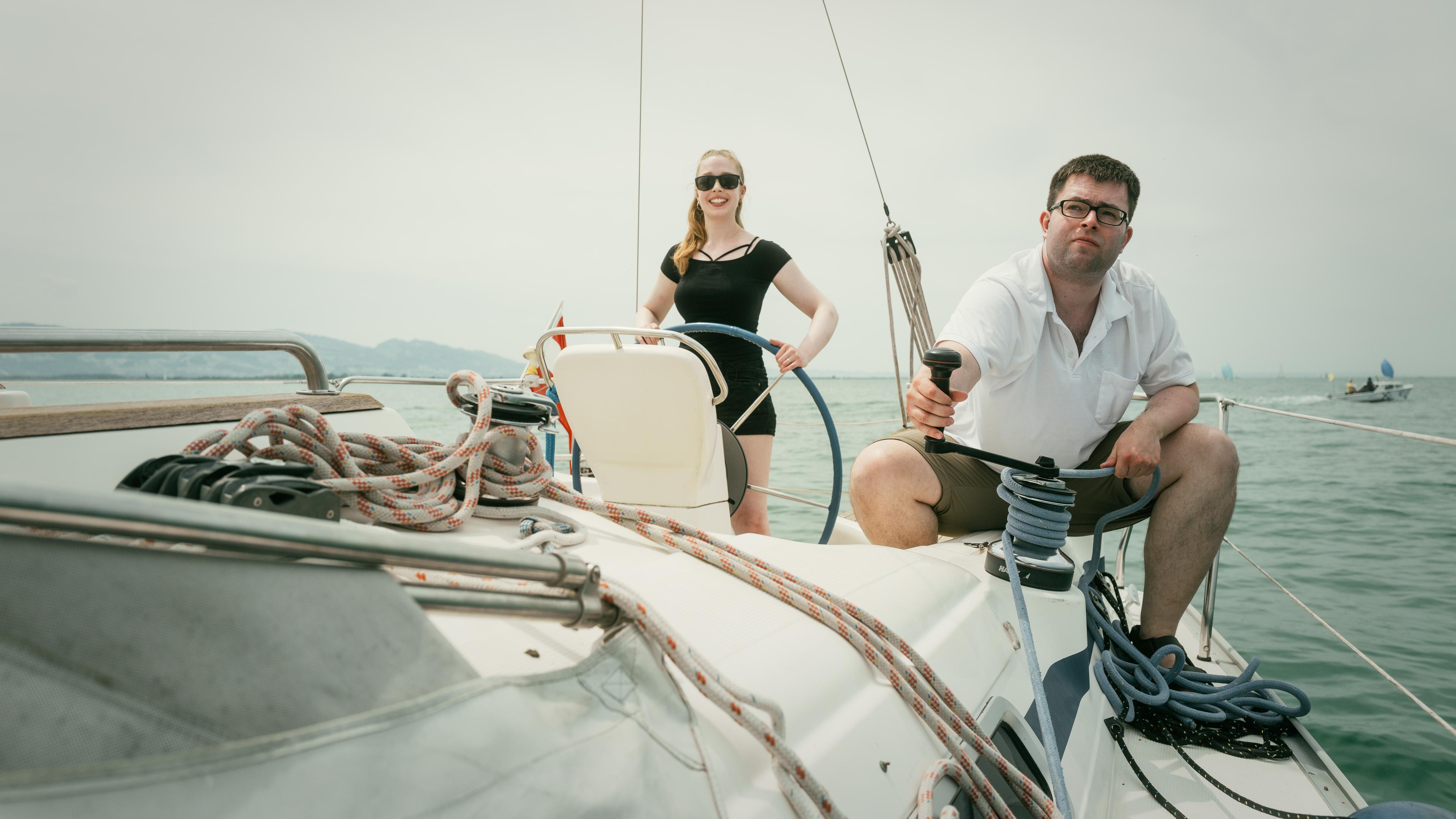 Christina steht am Heck des Segelboots, Christian sitzt in der Hocke an der Reling und hält das Tau des Segels.