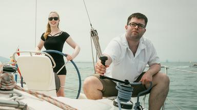 Christina steht am Heck des Segelboots, Christian sitzt in der Hocke an der Reling und hält das Tau des Segels.