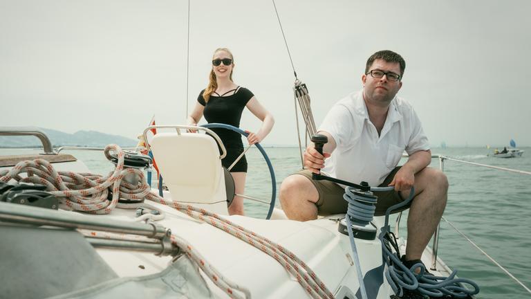 Christina steht am Heck des Segelboots, Christian sitzt in der Hocke an der Reling und hält das Tau des Segels.