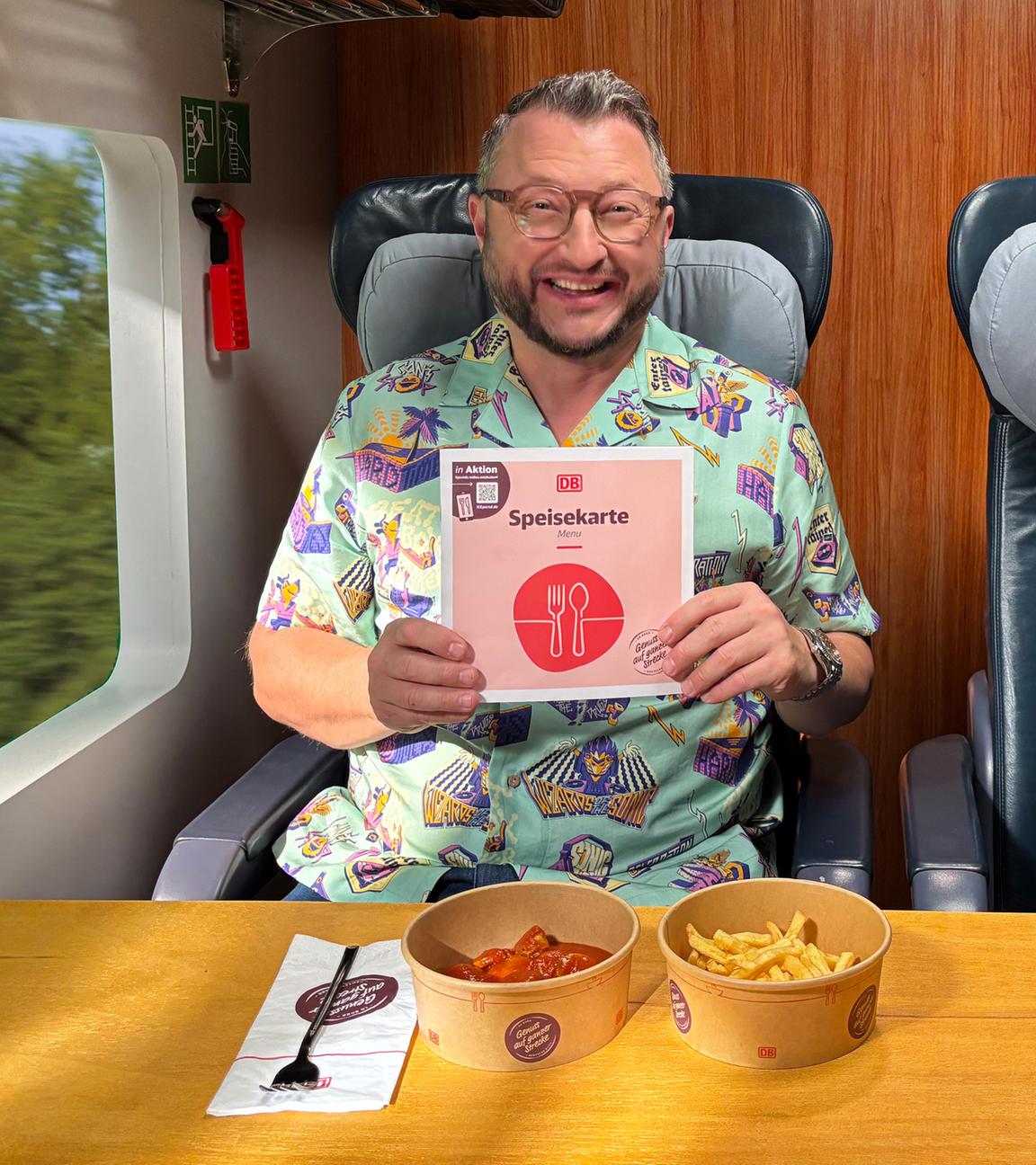 Mann im Zug mit buntem Hemd hält eine Speisekarte des DB-Bordbistros in der Hand; vor ihm stehen Currywurst mit Pommes und ein Einwegbecher für Heißgetränke.