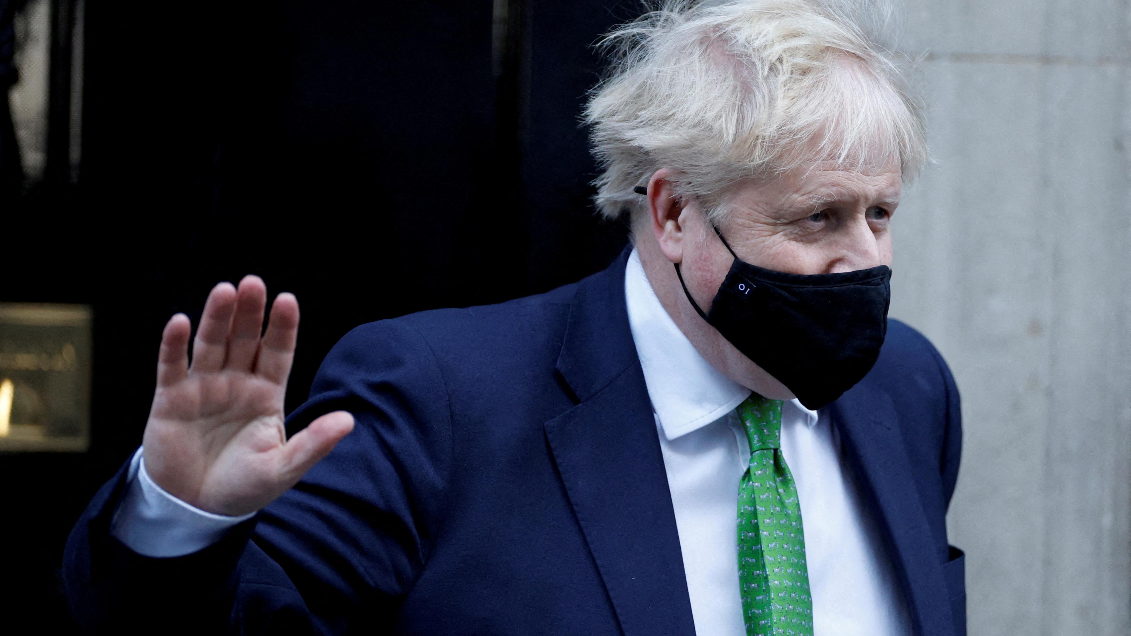 Boris Johnson mit schwarzer Maske und grüner Krawatte hebt die Hand vor Downing Street 10 in London
