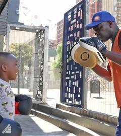 Boxtrainer George Khosi trägt ein orangenes Hemd und hat an den Händen jeweils ein Schlagpolster. Ihm gegenüber stehen drei Kinder mit Boxhandschuhen an den Händen.