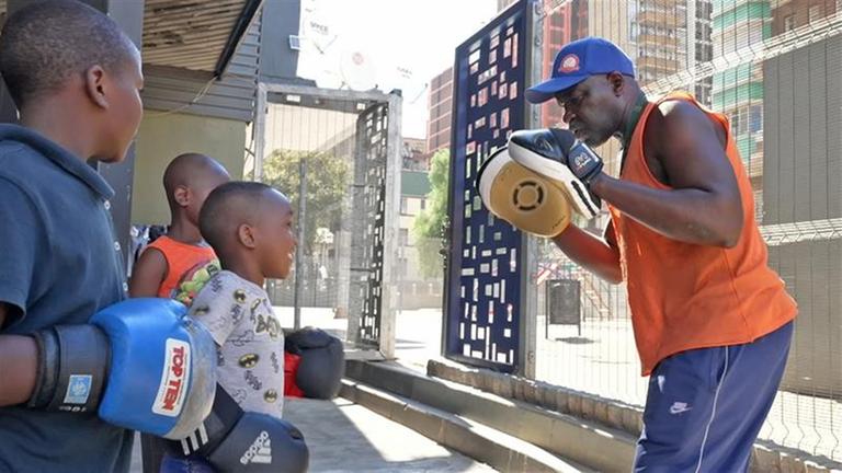 Boxtrainer George Khosi trägt ein orangenes Hemd und hat an den Händen jeweils ein Schlagpolster. Ihm gegenüber stehen drei Kinder mit Boxhandschuhen an den Händen.