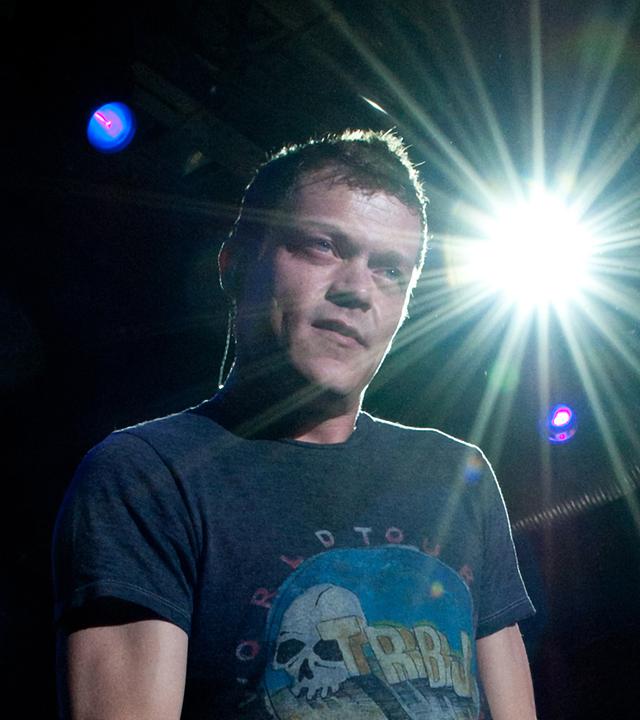 Der Sänger Brad Arnold steht auf der Bühne, von hinten strahlt Scheinwerferlicht in die Kamera.