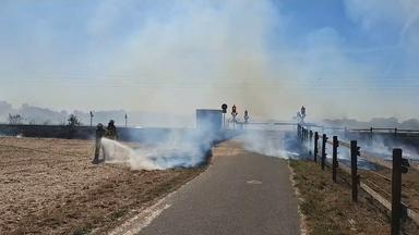 Feuerwehrkräfte löschen einen Böschungsbrand entlang einer Bahnstrecke. Rauch breitet sich über die Fläche aus, neben den Gleisen verläuft ein asphaltierter Weg.