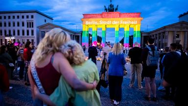 Menschen vor dem Brandenburger Tor, dass in Regenbogenfarben leuchtet. Darauf ist der Schriftzug zu lesen: "Berlin, Stadt der Freiheit"