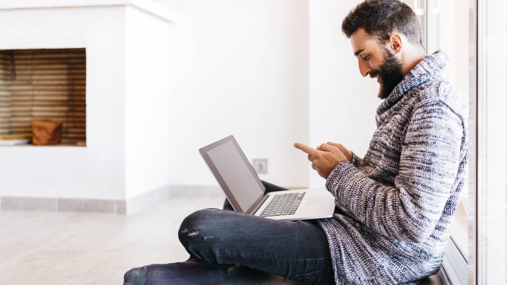 Ein Mann sitzt mit einem Laptop und einem Smartphone auf dem Boden vor einem Kamin. 