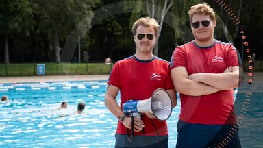 Pablo Spallek und sein Kollege Jakob stehen am Beckenrand eines Freibads. Beide tragen rote T-Shirts und blaue Shorts mit dem Logo der "Berliner Bäder" sowie eine Sonnenbrille. Pablo Spallek hält ein Megafon in der Hand. Jakob hat die Arme vor der Brust verschränkt. Im Hintergrund schwimmen Menschen im Wasser.