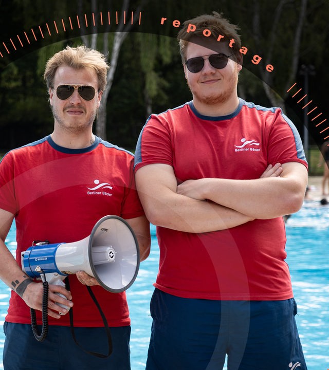 Pablo Spallek und sein Kollege Jakob stehen am Beckenrand eines Freibads. Beide tragen rote T-Shirts und blaue Shorts mit dem Logo der "Berliner Bäder" sowie eine Sonnenbrille. Pablo Spallek, hält ein Megafon in der Hand. Jakob, hat die Arme vor der Brust verschränkt. Im Hintergrund schwimmen Menschen im Wasser.