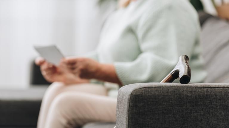 Eine ältere Frau sitzt mit Gehstock auf dem Sofa und liest einen Brief.