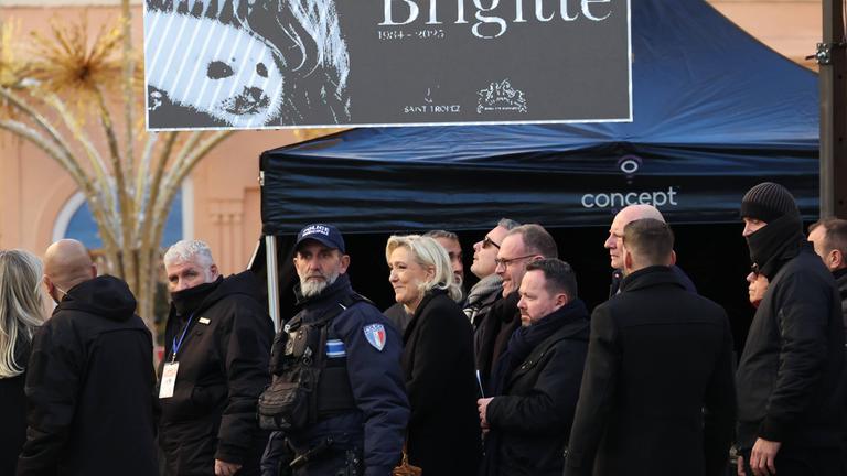 Mehrere Personen, darunter Marine Le Pen sowie Polizisten, in überwiegend schwarzer Kleidung. Über ihnen eine Art Plakat mit der Aufschrift "Brigitte".