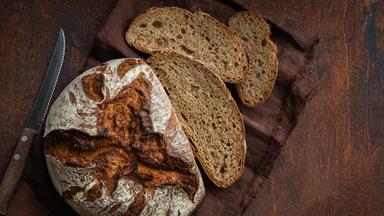 Ein frisch gebackener Laib Brot liegt auf dem Tisch, links daneben liegen ein Messer und rechts mehrere geschnittene Brotscheiben.