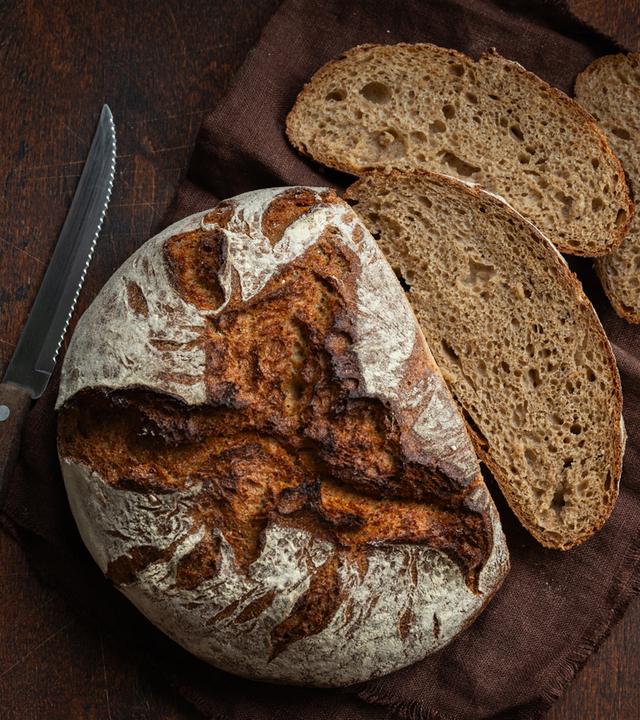 Ein frisch gebackener Laib Brot liegt auf dem Tisch, links daneben liegen ein Messer und rechts mehrere geschnittene Brotscheiben.
