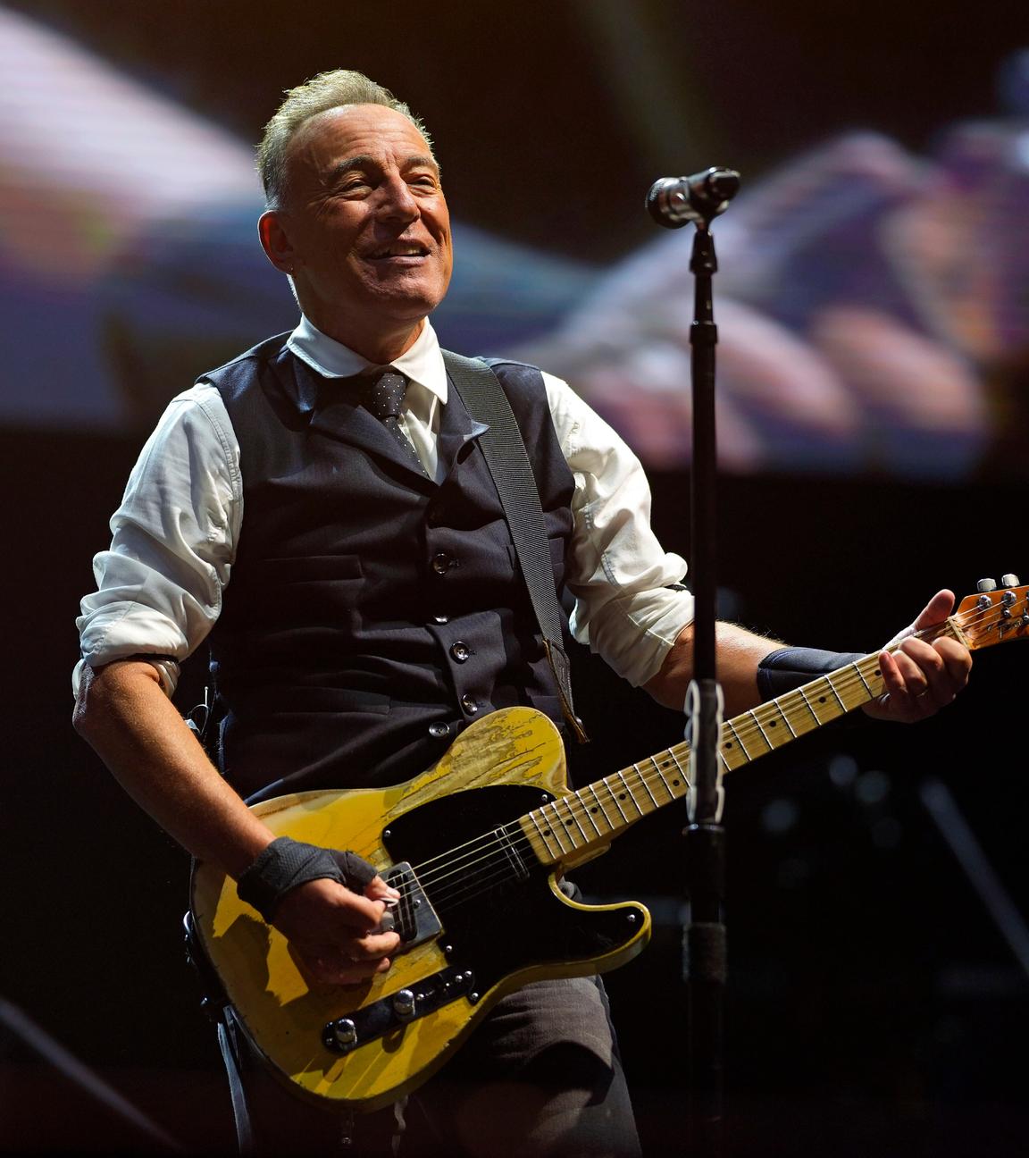 Der Musiker Bruce Springsteen. Er steft auf der Bühne, hält eine Gitarre und singt in ein Mikrofon.