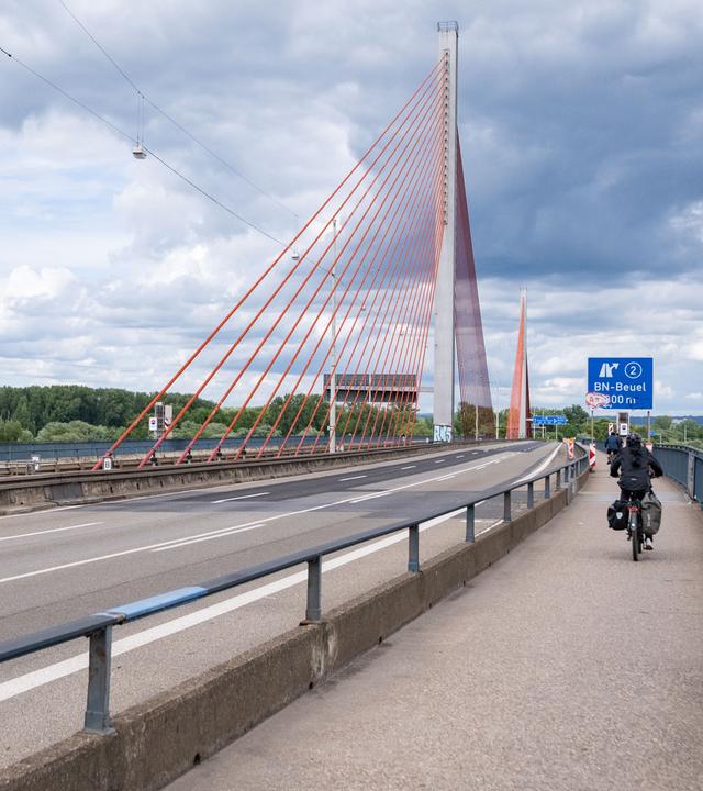 Auf dem Bild sieht man die Bonner Rheinbrücke A565. Die Fahrbahn ist leer und rechts auf dem Bürgersteig rechts fahren zwei Fahrradfahrer:innen.