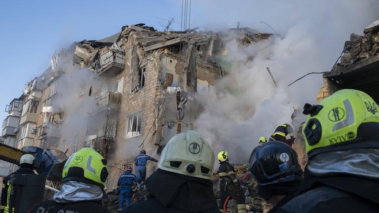 Im Hintergrund ist ein verschüttetes Gebäude aus dem Rauch kommt. Im Vordergrund sind Feuerwehrmänner