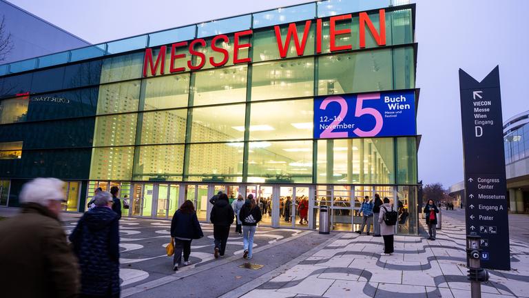 Menschen laufen über einen Platz in einen großen modernen Glasbau, auf dem Messe Wien steht und ein Plakat der Buch Wien 25 hängt.