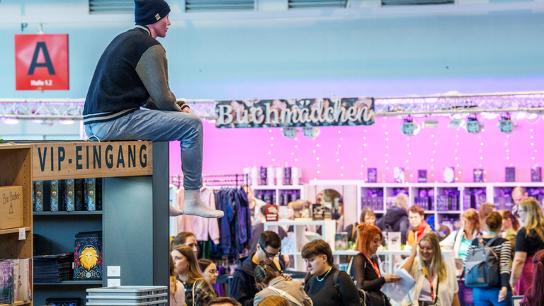 Ein Mann sitzt oben auf einer Stellwand zwischen Buchmessen-Gängen und Ständen über dem Menschengewusel.