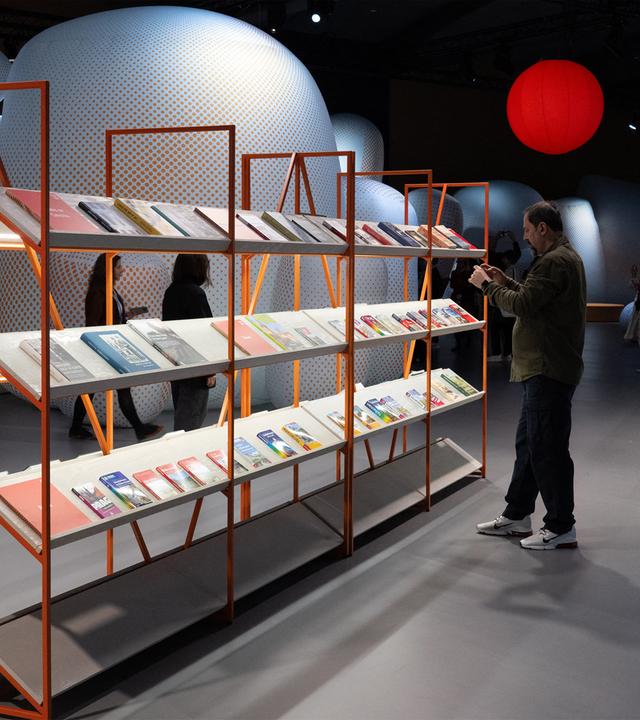 In einer abgedunkelten Halle steht ein filigranes Bücherregal aus Holz, vor dem ein Mann ein Buch liest. Im Hintergrund sind weitere Besucher zu sehen und runde futuristisch anmutende Objekte sowie eine rot leuchtende Kugel.