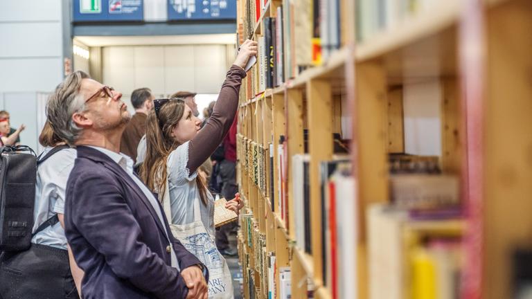 Vor einem großen Bücherregal stehen Menschen und betrachten die Buchrücken, oder ziehen Bücher heraus.