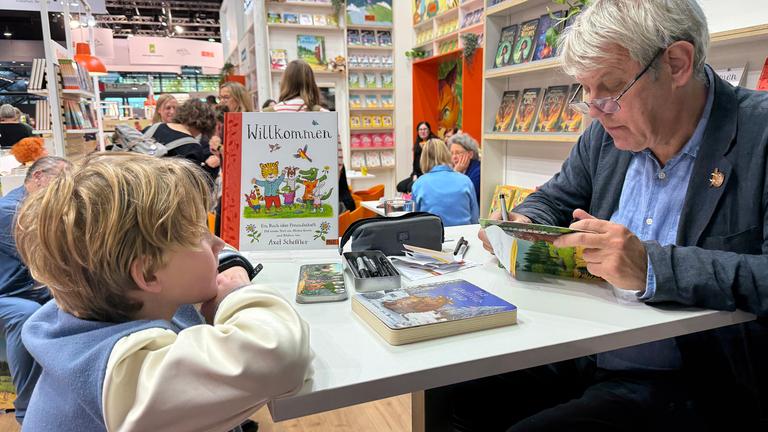 Ein Mann sitzt an einem Büchertisch und hält ein Buch in den Händen, ihm gegenüber leht sich ein kleiner Junge auf den Tisch und sieht ihm zu. Beide sind umgeben von Stellwänden, auf denen Bücher präsentiert werden.