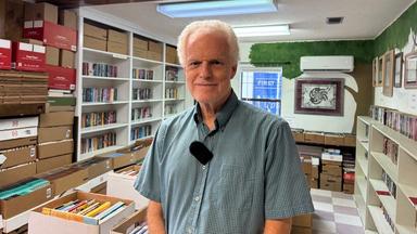 Ein Mann in einem kurzen, karierten Hemd steht in einem Raum voller Bücher und schaut freundlich in die Kamera.
