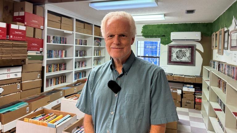 Ein Mann in einem kurzen, karierten Hemd steht in einem Raum voller Bücher und schaut freundlich in die Kamera.