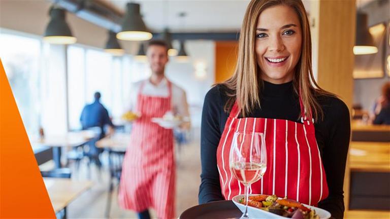Bei Teilzeit-Servicekräften erfolgt oft eine Aufstockung: Frau trägt Tablett mit Essen und einem Glas Wein in einem Restaurant