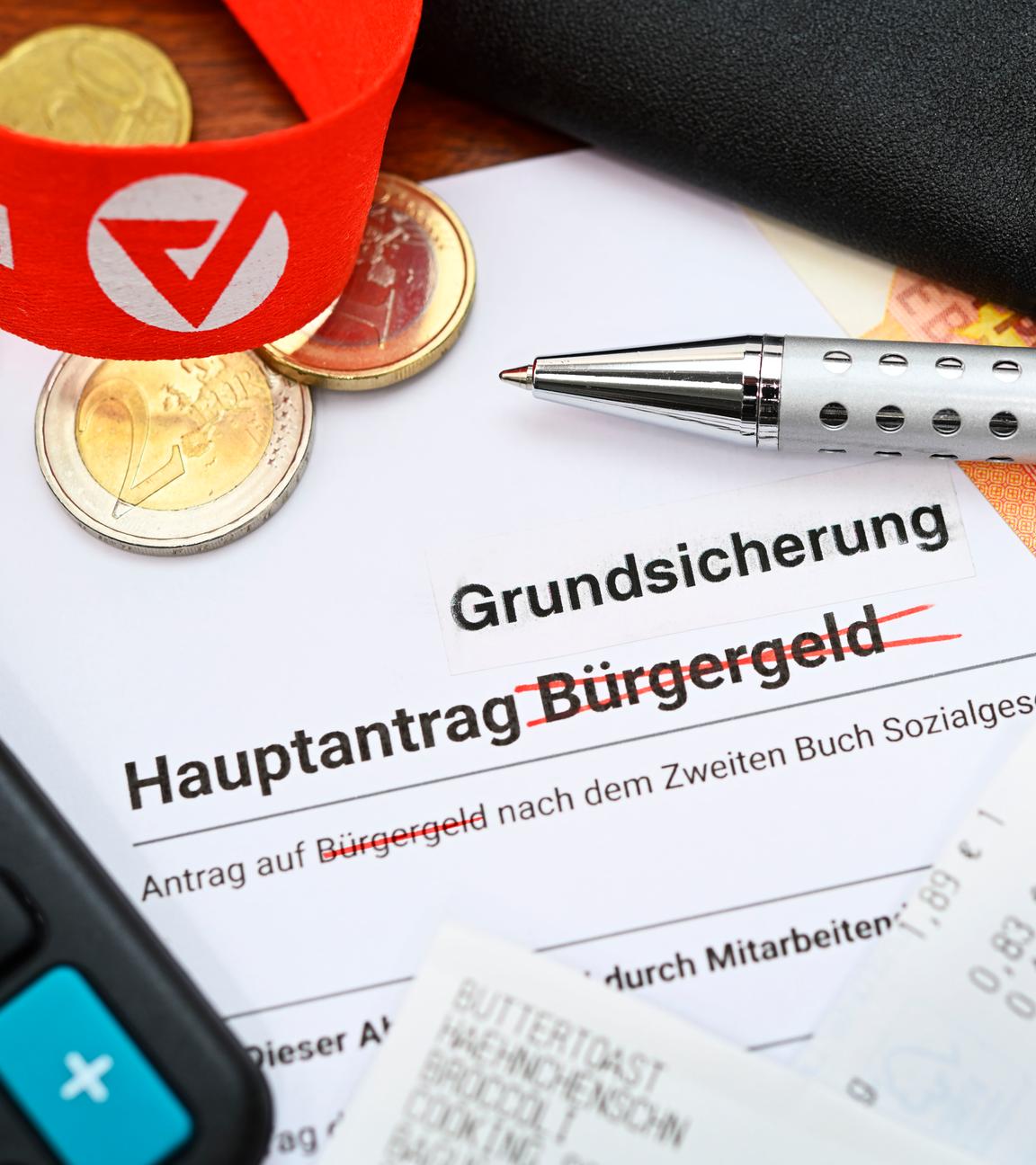 Ein Antrag, auf dem das Wort "Bürgergeld" durchgestrichen und "Grundsicherung" oben drüber geschrieben wurde. Darum herum Bargeld, ein Taschenrechner und das Logo der Agentur für Arbeit auf einem Schlüsselband.