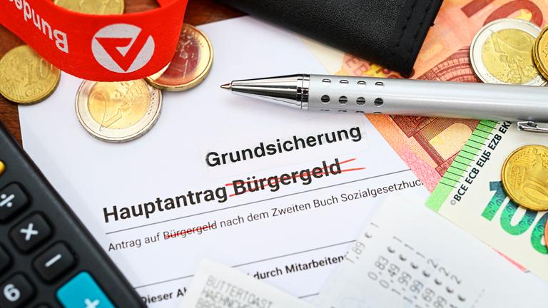 Ein Antrag, auf dem das Wort "Bürgergeld" durchgestrichen und "Grundsicherung" oben drüber geschrieben wurde. Darum herum Bargeld, ein Taschenrechner und das Logo der Agentur für Arbeit auf einem Schlüsselband.