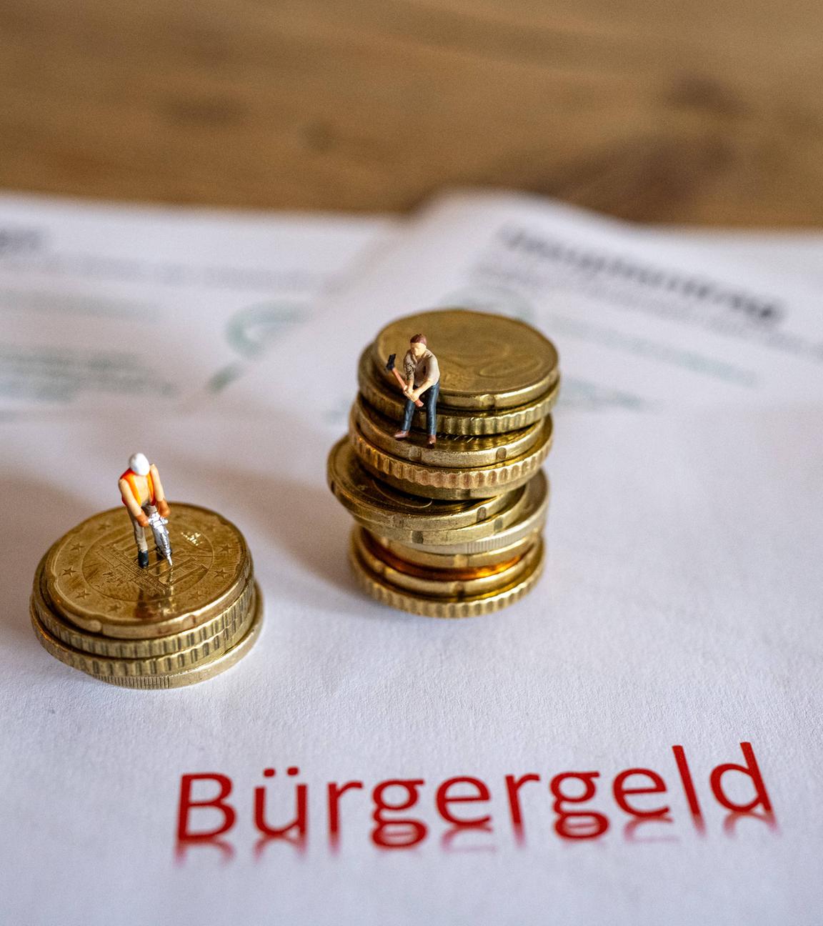 Das Bild zeigt symbolische Antragsformulare für das Bürgergeld sowie Geldmünzen und symbolische Spielzeugfiguren in Form von Bauarbeitern.