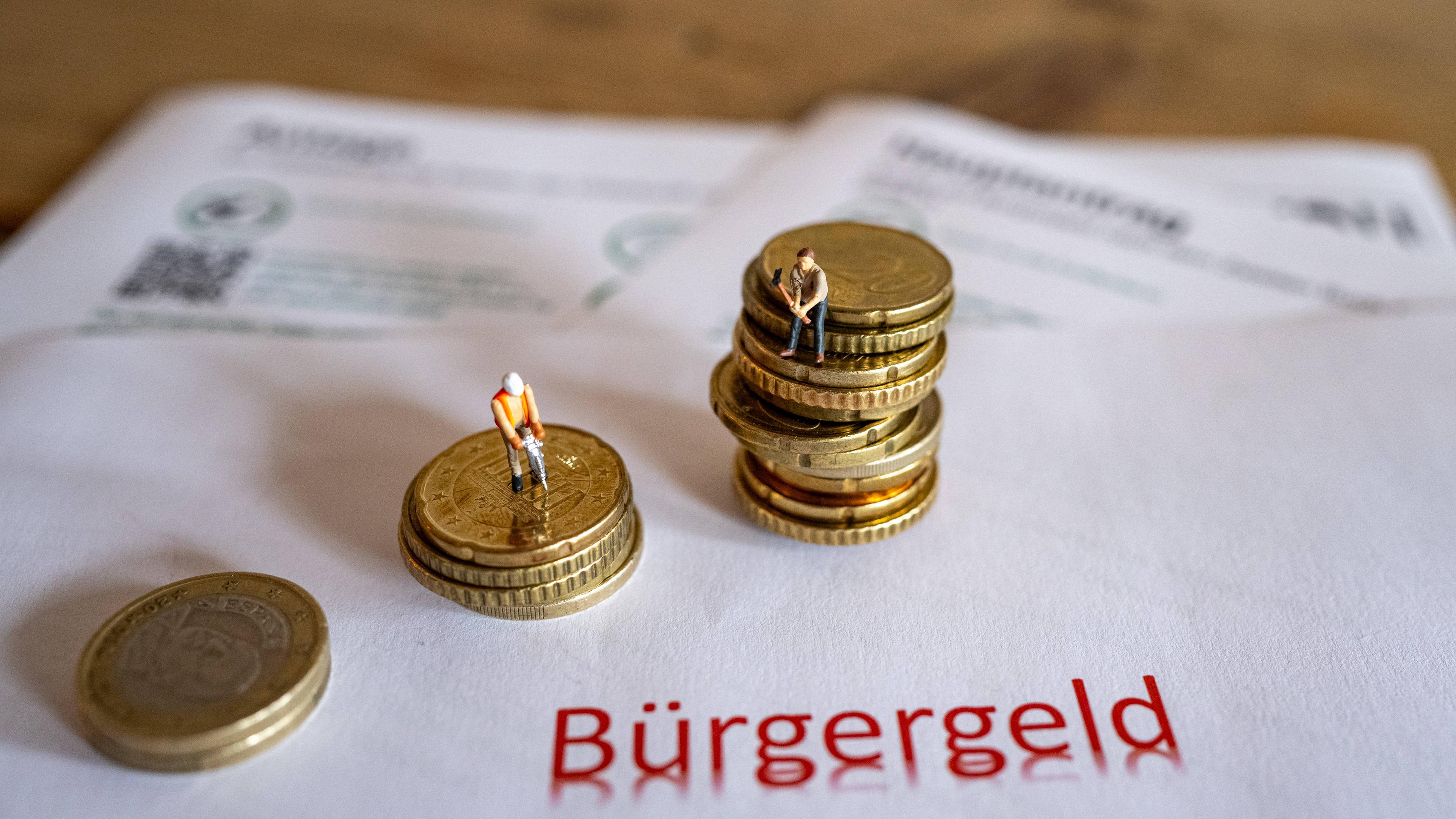 Das Bild zeigt symbolische Antragsformulare für das Bürgergeld sowie Geldmünzen und symbolische Spielzeugfiguren in Form von Bauarbeitern.