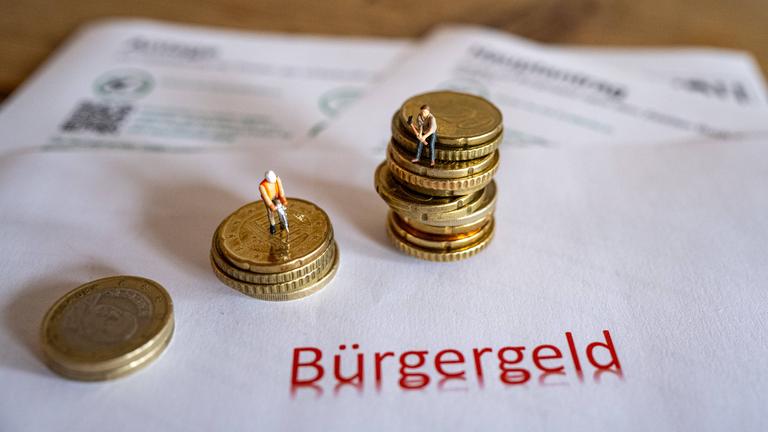 Das Bild zeigt symbolische Antragsformulare für das Bürgergeld sowie Geldmünzen und symbolische Spielzeugfiguren in Form von Bauarbeitern.