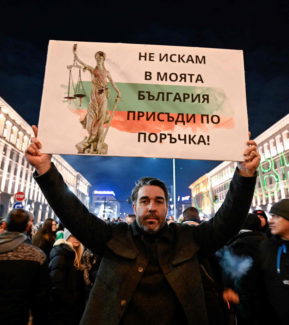 Ein Mann inmitten einer Masse von Demonstranten hält ein Plakat hoch. Auf diesem sind die Farben Bulgariens, ein Text in bulgarischer Schrift und die Justicia zu sehen.