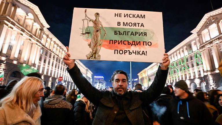 Ein Mann inmitten einer Masse von Demonstranten hält ein Plakat hoch. Auf diesem sind die Farben Bulgariens, ein Text in bulgarischer Schrift und die Justicia zu sehen.