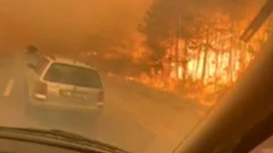 Ein Auto steht auf einer Autobahn, direkt neben einem Waldbrand, der alles in rot-oranges Licht taucht.