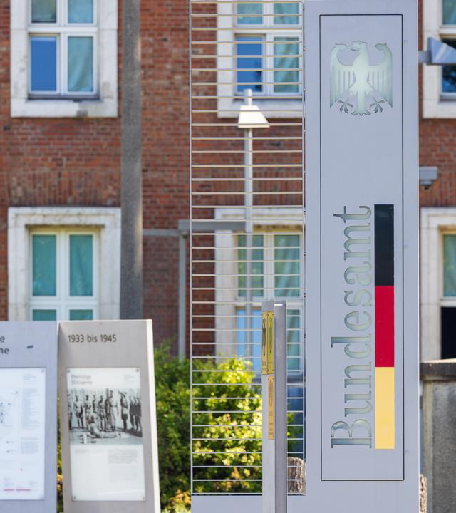Eine Stele mit dem Bundesadler und der Aufschrift "Bundesamt" in den Farben der deutschen Flagge markiert den Eingang zu einer Behörde.