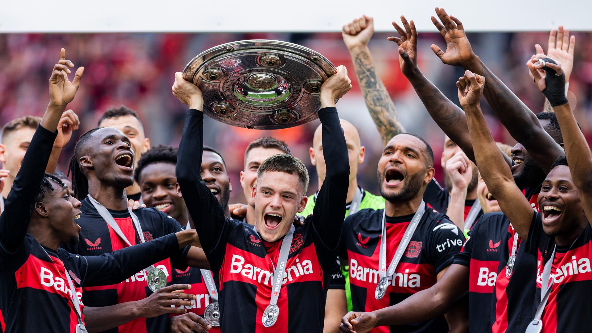 Florian Wirtz streckt die Meisterschale, umjubelkt von seinen Mannschaftskollegen des Bayer 04 Leverkusens, hoch.