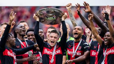 Florian Wirtz streckt die Meisterschale, umjubelkt von seinen Mannschaftskollegen des Bayer 04 Leverkusens, hoch.