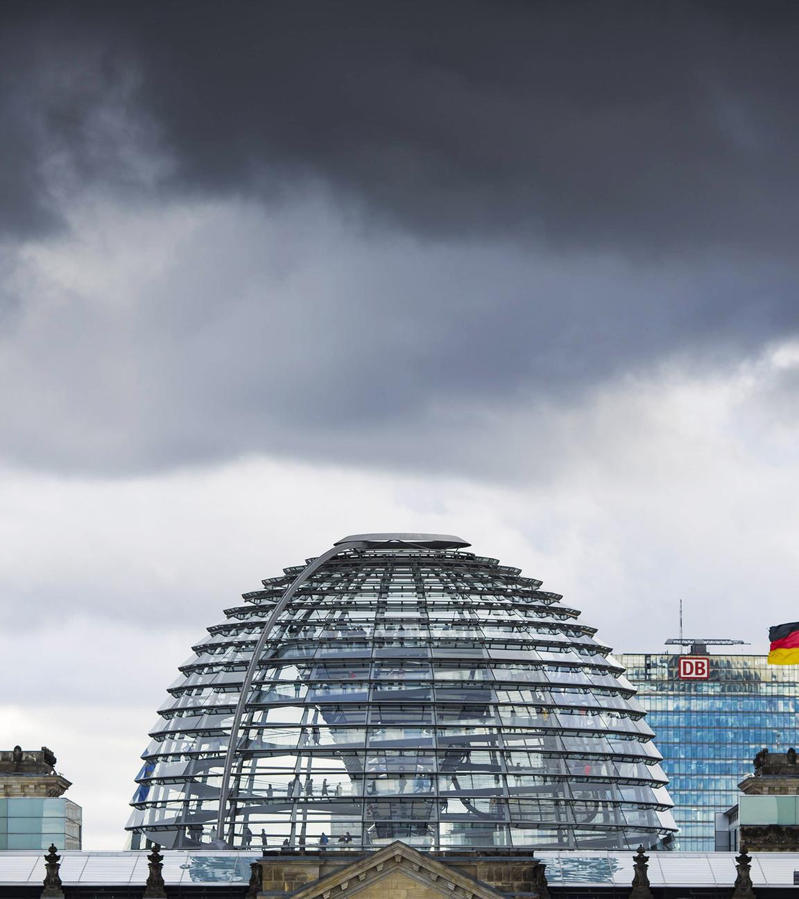 Über dem Bundestag mit seiner gläsernen Kuppel hängen dunkle Wolken. Über jedem der vier alten Türme wehen die deutsche und europäische Flagge.