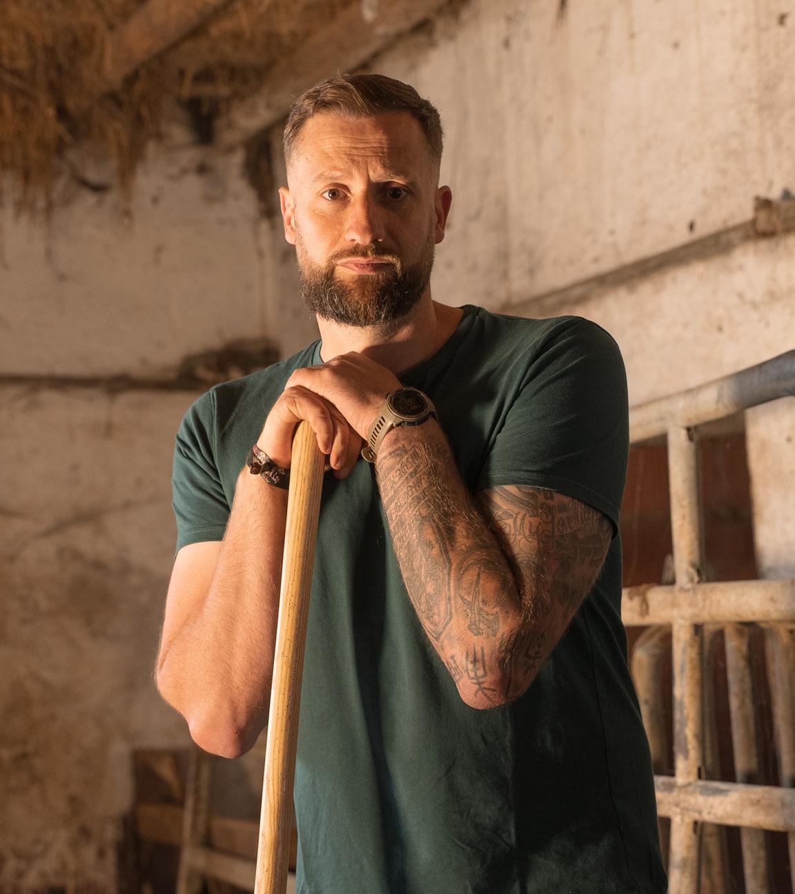 Christoph Rothhaupt, ein Mann mit braunen Haaren und Bart, steht in einem Stall. Er stützt auf einer Mistgabel und schaut mit traurigem Blick in die Kamera. Sein linker Arm ist tätowiert, er trägt eine Uhr. Im Hintergrund ist eine verschmutzte Wand und eine Decke mit Holzbalken, zwischen denen Stroh raus ragt.