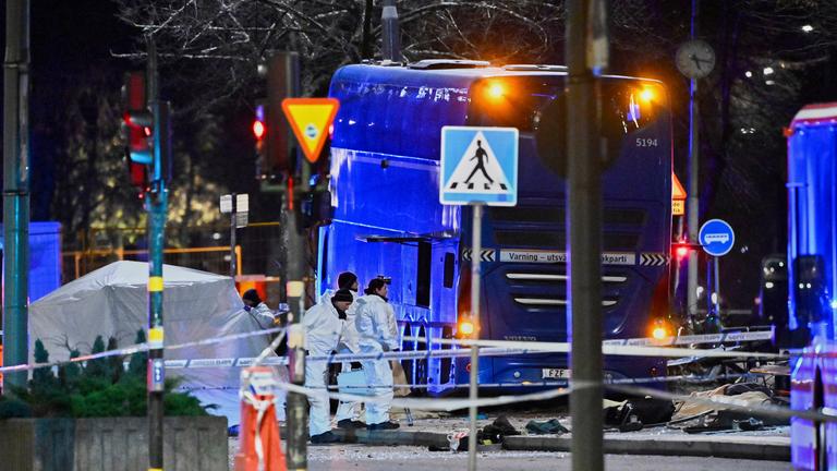 Einsatzkräfte in Schutzanzügen stehen an der Unfallstelle, an der ein Doppeldeckerbus in ein Wartehäuschen krachte und dabei Tote und Verletzte am 14.11.2025 im schwedischen Stockholm forderte.