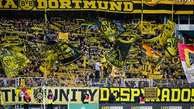 Fans und Ultras des BVBs schwingen geld-schwarze Borussia-Dortmund-Fahnen auf der Tribühne eines Stations. 