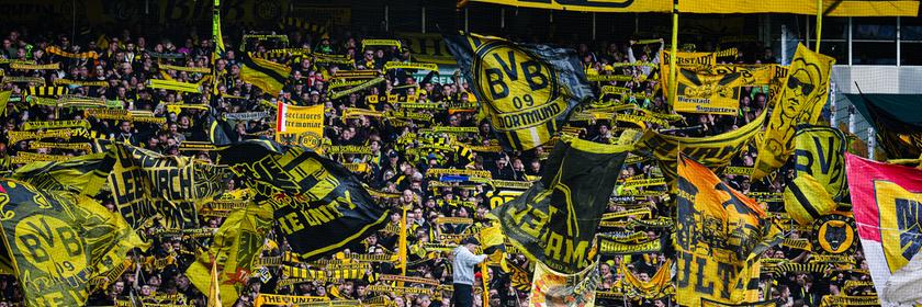 Fans und Ultras des BVBs schwingen geld-schwarze Borussia-Dortmund-Fahnen auf der Tribühne eines Stations. 