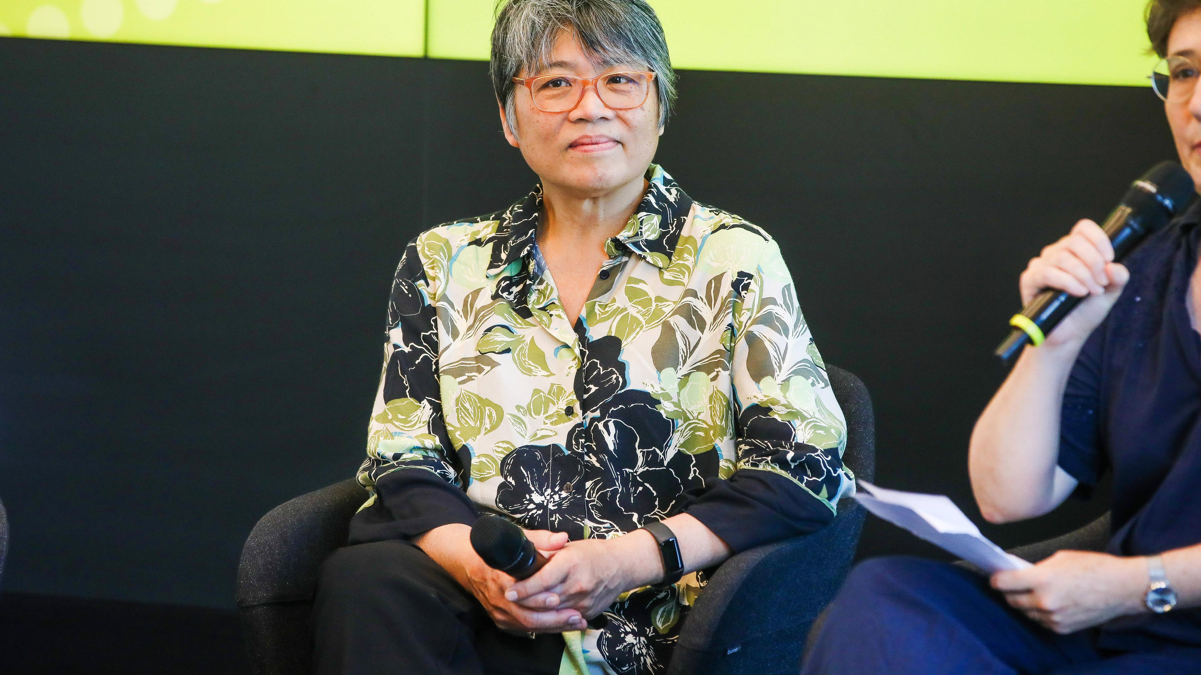 Foto einer asiatisch aussehenden Frau mittleren Alters mit kurzen graumelierten Haaren und Brille, die ein grün, weiß, grau gemustertes Hemd trägt.