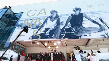 Vor dem Eingang zum Festivalkino in Cannes steht eine Gruppe Menschen. Über ihnen ist das riesige Plakat des Filmfestivals Cannes zu sehen.