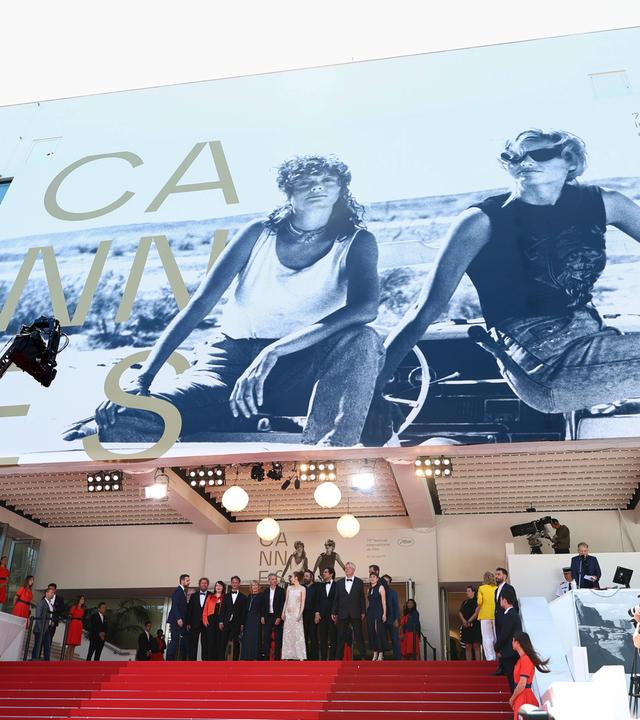 Vor dem Eingang zum Festivalkino in Cannes steht eine Gruppe Menschen. Über ihnen ist das riesige Plakat des Filmfestivals Cannes zu sehen.